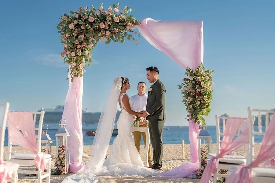 bride and groom on beach under an arch with pink flowers. The ocean in the background